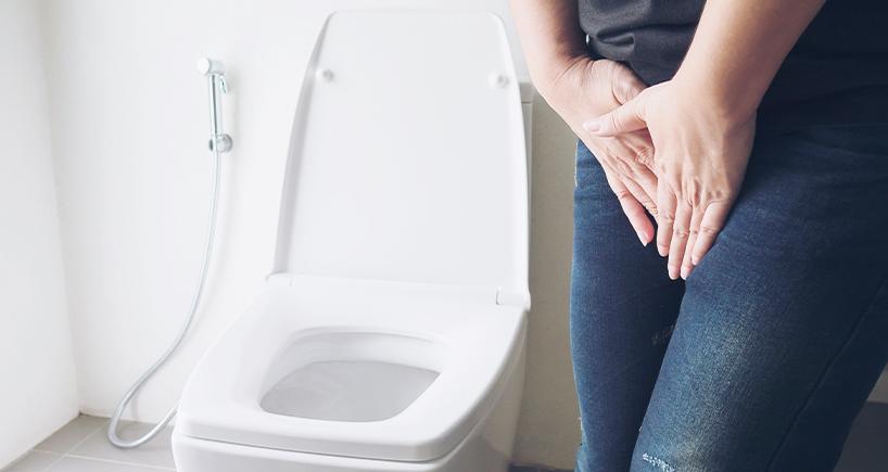 A person holding their hands near their crotch stands beside an open toilet, suggesting a need to urinate.